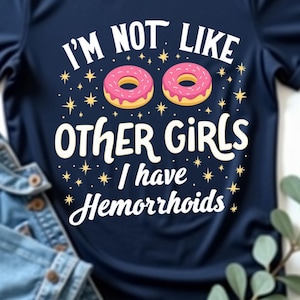 Puede incluir: Camiseta azul marino con el texto "I'M NOT LIKE OTHER GIRLS I have Hemorrhoids" y dos donuts rosas. Una chaqueta vaquera está al lado de la camiseta. La camiseta es una prenda informal.