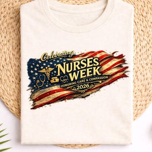 May include: A cream-colored t-shirt with a graphic celebrating Nurses Week 2026. The design features an American flag with a medical symbol and the words "Nurses Week" in gold lettering. The shirt is laid flat on a woven mat.