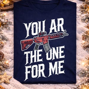 Op de afbeelding: Marineblauw t-shirt met de tekst "YOU ARE THE ONE FOR ME" en een rode geweer afbeelding. De afbeelding wordt gepresenteerd tegen een feestelijke achtergrond met lichtjes en ornamenten.