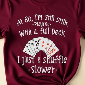 Pode incluir: Camiseta bordô com a frase "At 80, I'm still playing with a full deck, I just shuffle slower" e um gráfico de cartas de baralho. A camiseta é amarrada na parte inferior.