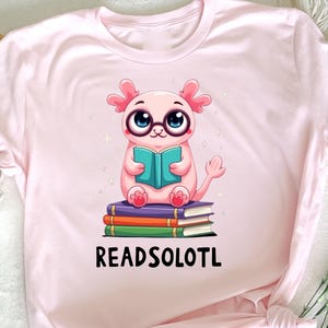 May include: Pink t-shirt with a cartoon axolotl wearing glasses and reading a book. The axolotl is sitting on a stack of books. The text "READSOLOTL" is printed below the axolotl.