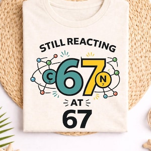 May include: Beige t-shirt with the text "STILL REACTING AT 67". The graphic features a stylized molecular structure with the numbers 6 and 7 in teal and yellow. The shirt is laid on a woven mat with a straw hat and other decorative items.