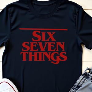 May include: A black t-shirt with the red text "SIX SEVEN THINGS". The image also shows light blue jeans and white trainers. The word "BLACK" is in the top left corner.