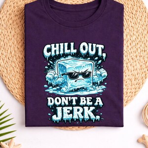 May include: A dark purple t-shirt with the words "CHILL OUT, DON'T BE A JERK" and a cartoon ice cube wearing sunglasses. The shirt is displayed on a woven mat with decorative items.