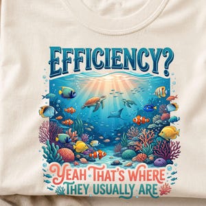 May include: A natural-coloured t-shirt featuring a graphic of an underwater coral reef scene with various colourful fish, a sea turtle, and a manta ray. The text EFFICIENCY? is printed in blue above, and YEAH THAT'S WHERE THEY USUALLY ARE below.
