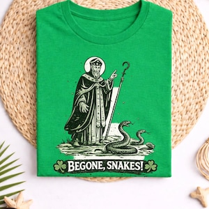 May include: Irish green t-shirt featuring a graphic of a saint, snakes, and the text "BEGONE, SNAKES!". The saint is shown with a staff, and the snakes are coiled. The shirt is displayed on a woven mat.