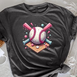 May include: Dark grey heather t-shirt with a graphic of a baseball with pink and blue bats crossed behind it. The baseball is on a pink and white base with pink and blue sprinkles around it.