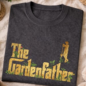 May include: Dark grey heather crew neck t-shirt with the text "The Gardenfather" in a gold, textured font with green floral accents. A gold silhouette of a person gardening is on the right side of the text.