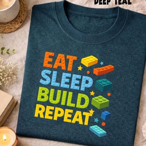 May include: Deep teal t-shirt with the words "EAT SLEEP BUILD REPEAT" in orange, blue, green, and yellow block letters. The shirt also features colourful block graphics.