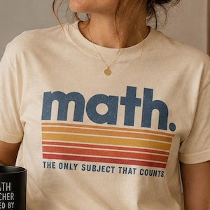 May include: A cream-colored short-sleeve t-shirt featuring the word math. in blue text above horizontal stripes in yellow, orange, and red, with the text THE ONLY SUBJECT THAT COUNTS. A black mug displays the text MATH TEACHER FUELED BY COFFEE & INTEGRALS.