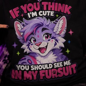 Puede incluir: Una camiseta negra con el gráfico de un personaje felino antropomórfico de color morado y blanco con ojos grandes. El texto dice IF YOU THINK I'M CUTE YOU SHOULD SEE ME IN MY FURSUIT, decorado con estrellas coloridas y un icono de huella.