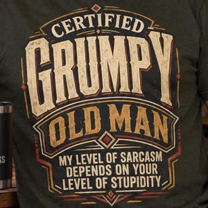 Może przedstawiać: Oliwkowozielony t-shirt z napisem CERTIFIED GRUMPY OLD MAN oraz MY LEVEL OF SARCASM DEPENDS ON YOUR LEVEL OF STUPIDITY w czcionce w stylu vintage. Czarny kubek podróżny z napisem BUILT FOR EARLY MORNINGS jest trzymany w jednej dłoni.