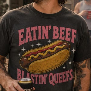 May include: A charcoal grey short-sleeve t-shirt featuring a graphic of a hot dog with mustard and the text EATIN' BEEF BLASTIN' QUEEFS in pink lettering, accented by small white star shapes surrounding the central hot dog illustration.