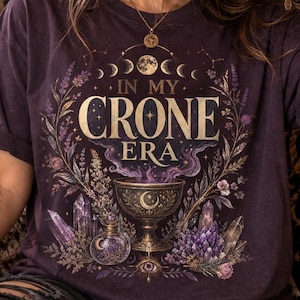 May include: A dark purple short-sleeve t-shirt featuring a graphic print with the text "IN MY CRONE ERA" in gold lettering. The design includes a central chalice, purple crystals, botanical elements, and moon phases arranged in a circular pattern.