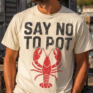 May include: A cream-colored short-sleeve t-shirt featuring the black distressed text SAY NO TO POT above a red graphic illustration of a lobster. The shirt has a crew neckline and a standard fit, showcasing a vintage-inspired graphic design.