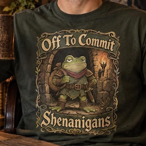 Puede incluir: Una camiseta de color verde oliva oscuro con un gráfico de una rana vestida de aventurero de pie en un arco de piedra. El texto encima y debajo de la rana dice "Off To Commit" y "SHENANIGANS" en una fuente decorativa de estilo vintage.