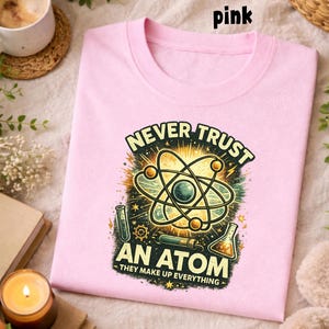 Puede incluir: Una camiseta rosa claro con cuello redondo. La camiseta presenta un gr&aacute;fico de un &aacute;tomo con el texto "NEVER TRUST AN ATOM - THEY MAKE UP EVERYTHING" en un estilo vintage. La palabra "pink" est&aacute; en la parte superior.