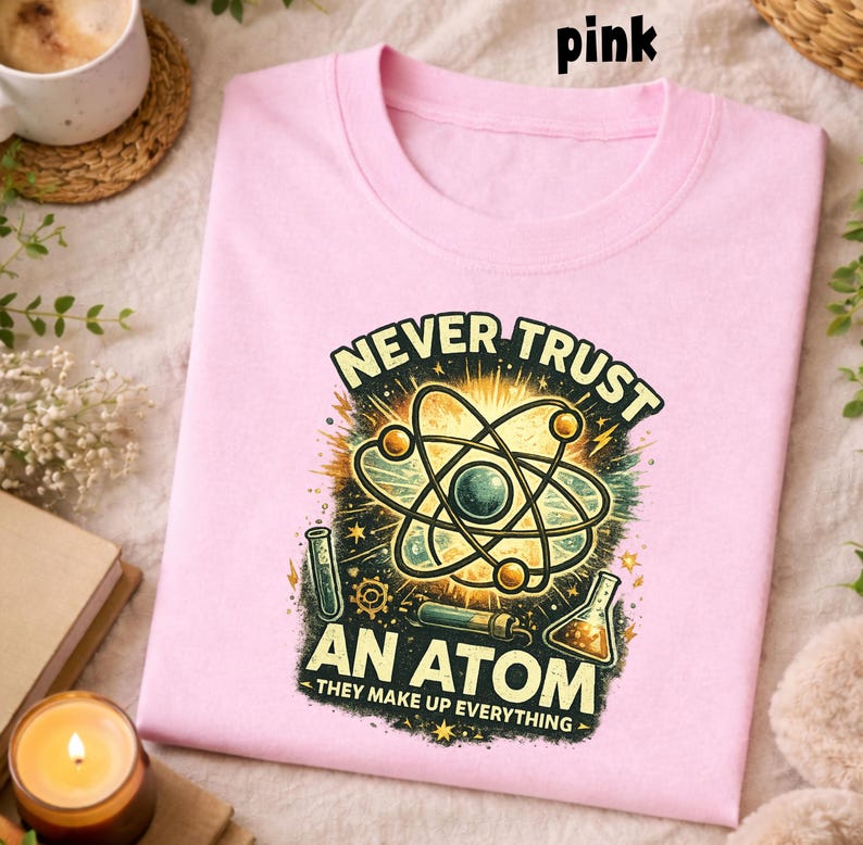 Puede incluir: Una camiseta rosa claro con cuello redondo. La camiseta presenta un gr&aacute;fico de un &aacute;tomo con el texto "NEVER TRUST AN ATOM - THEY MAKE UP EVERYTHING" en un estilo vintage. La palabra "pink" est&aacute; en la parte superior.