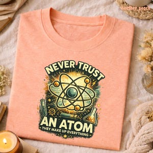 Puede incluir: Una camiseta de color melocot&oacute;n con un gr&aacute;fico de tem&aacute;tica cient&iacute;fica. El gr&aacute;fico presenta un modelo de &aacute;tomo, tubos de ensayo y el texto "NEVER TRUST AN ATOM - THEY MAKE UP EVERYTHING". La camiseta est&aacute; doblada.