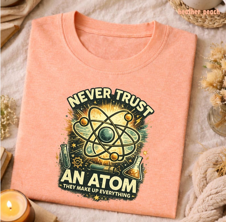 Puede incluir: Una camiseta de color melocot&oacute;n con un gr&aacute;fico de tem&aacute;tica cient&iacute;fica. El gr&aacute;fico presenta un modelo de &aacute;tomo, tubos de ensayo y el texto "NEVER TRUST AN ATOM - THEY MAKE UP EVERYTHING". La camiseta est&aacute; doblada.