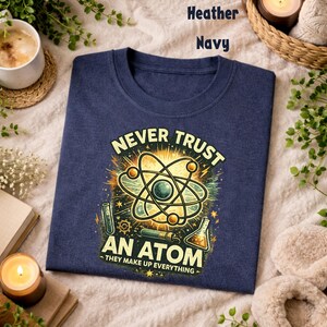 Puede incluir: Camiseta azul marino con un gr&aacute;fico de tem&aacute;tica cient&iacute;fica. El dise&ntilde;o presenta una ilustraci&oacute;n de un &aacute;tomo con el texto "NEVER TRUST AN ATOM - THEY MAKE UP EVERYTHING". El gr&aacute;fico es en tonos amarillos, verdes y azules.