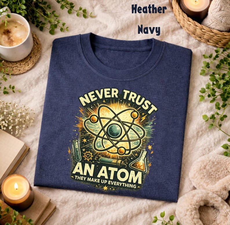 Puede incluir: Camiseta azul marino con un gr&aacute;fico de tem&aacute;tica cient&iacute;fica. El dise&ntilde;o presenta una ilustraci&oacute;n de un &aacute;tomo con el texto "NEVER TRUST AN ATOM - THEY MAKE UP EVERYTHING". El gr&aacute;fico es en tonos amarillos, verdes y azules.