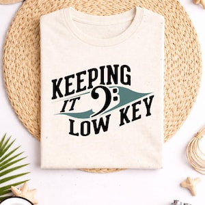 May include: Cream-colored t-shirt with the text "KEEPING IT LOW KEY" in black and teal. The design features a musical symbol. The shirt is laid flat on a woven mat, surrounded by decorative items.