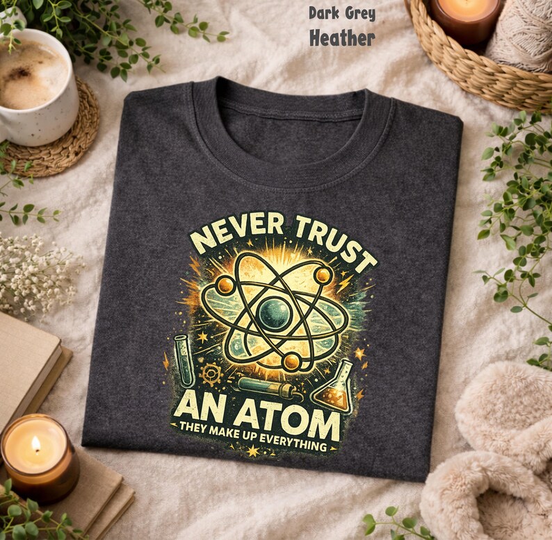 Puede incluir: Camiseta gris oscuro jaspeado con un dise&ntilde;o de tem&aacute;tica cient&iacute;fica. El dise&ntilde;o presenta un &aacute;tomo con el texto "NEVER TRUST AN ATOM - THEY MAKE UP EVERYTHING" en amarillo y verde.