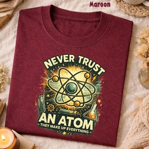 Puede incluir: Camiseta granate con un gr&aacute;fico de tem&aacute;tica cient&iacute;fica. El dise&ntilde;o presenta una ilustraci&oacute;n de un &aacute;tomo con el texto "NEVER TRUST AN ATOM - THEY MAKE UP EVERYTHING" en un estilo desgastado. La camiseta est&aacute; doblada y expuesta sobre una superficie neutra.