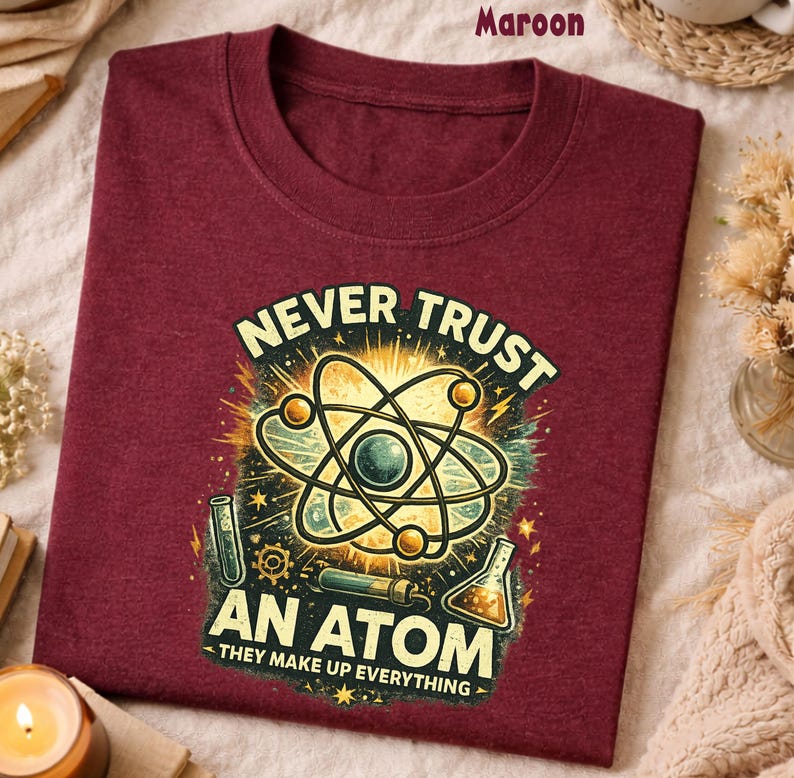 Puede incluir: Camiseta granate con un gr&aacute;fico de tem&aacute;tica cient&iacute;fica. El dise&ntilde;o presenta una ilustraci&oacute;n de un &aacute;tomo con el texto "NEVER TRUST AN ATOM - THEY MAKE UP EVERYTHING" en un estilo desgastado. La camiseta est&aacute; doblada y expuesta sobre una superficie neutra.