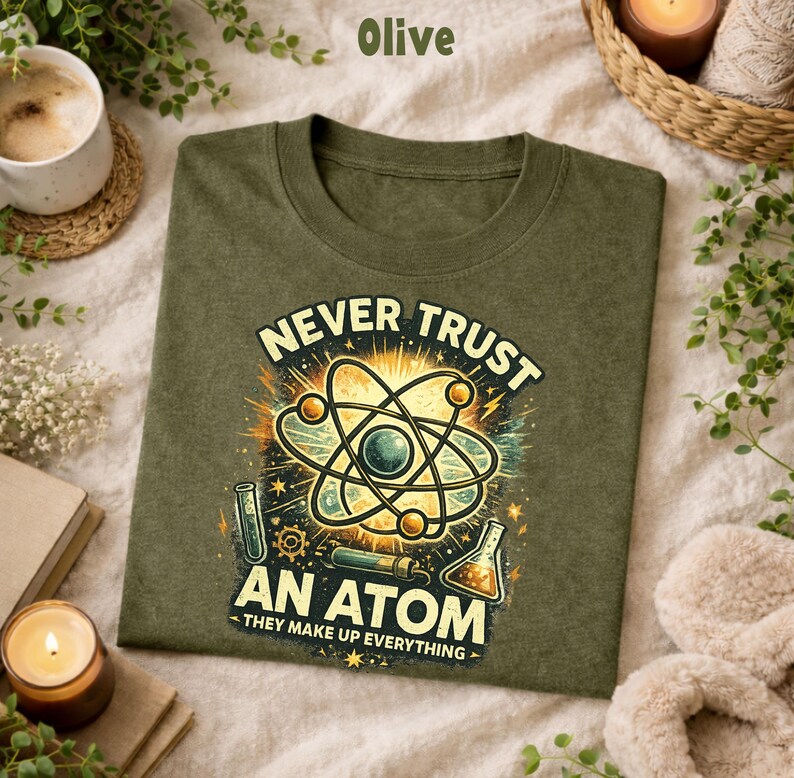 Puede incluir: Camiseta verde oliva con el texto "NEVER TRUST AN ATOM - THEY MAKE UP EVERYTHING" y un gr&aacute;fico de un &aacute;tomo. El gr&aacute;fico utiliza tonos amarillos, azules y verdes. La camiseta es de un material suave.