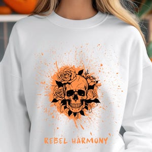 Może przedstawiać: Biała bluza z pomarańczowym wzorem czaszki otoczonej różami i liśćmi. Projekt zawiera napis "REBEL HARMONY" w kolorze pomarańczowym. Bluza ma okrągły dekolt i długie rękawy.