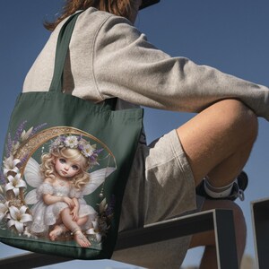 K&ouml;nnte beinhalten: Dunkelgr&uuml;ne Tragetasche mit einer Feenillustration. Die Fee hat blondes Haar, wei&szlig;e Fl&uuml;gel und ein wei&szlig;es Kleid, umgeben von wei&szlig;en Lilien, Lavendel und einem goldenen Halbmond. Die Tasche hat dunkelgr&uuml;ne Griffe.