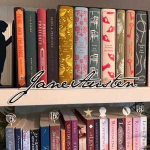May include: A bookshelf displaying a collection of Jane Austen novels, including "Pride and Prejudice" and "Sense and Sensibility." The books have decorative covers in various colors. A cursive "Jane Austen" sign sits in front of the books. Bookmarks and decorative objects are also visible.