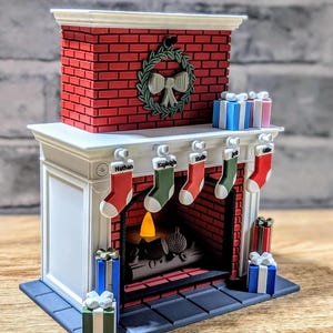 May include: A miniature fireplace model with a red brick facade, a white mantel, and a green wreath with a bow. Five stockings labelled Nathan, Kayleigh, Marie, Jack, and Ollie hang from the mantel. Wrapped gift boxes sit on the mantel and floor.