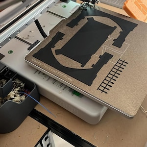 May include: A 3D printer with a white frame and a gold-coloured Bambu Textured PEI Plate. The print bed features a black plastic object in the process of being manufactured, with a grid-like structure and solid geometric shapes visible.