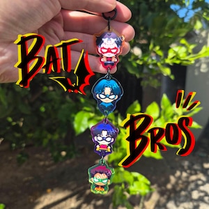 Puede incluir: Un llavero vertical de acrílico con cuatro personajes de superhéroes estilo chibi conectados por anillos de metal negro. Los personajes visten trajes rojos, azules, morados y verdes. El texto BAT BROS aparece en letras negritas rojas y negras.