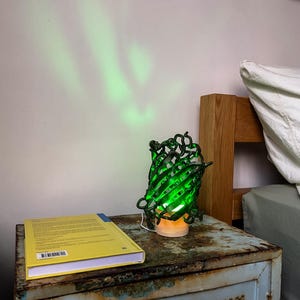 Può includere: Una lampada verde a filo metallico con interno luminoso è appoggiata su un comodino blu usurato. Un libro giallo con dorso blu è appoggiato accanto alla lampada. La lampada proietta una luce verde sulla parete. Il comodino è accanto a una struttura letto in legno.