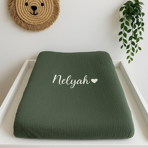 Peut inclure: Un matelas à langer vert olive avec le nom "Nelyah" en écriture blanche et un symbole de cœur. Une décoration murale en forme de tête de lion tressée et une plante suspendue sont visibles en arrière-plan.