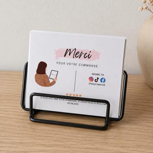 Peut inclure: Une carte de remerciement rectangulaire blanche avec du texte noir et un graphique représentant une personne tenant une tablette, présentée dans un support en fil métallique noir. La carte affiche le mot Merci, des icônes sociales et cinq étoiles.