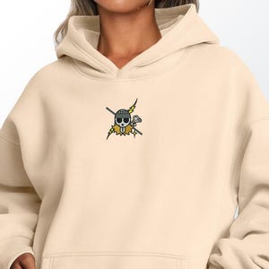 May include: Cream-colored hoodie with a front pocket and a hood. The hoodie features a small embroidered design of a skull with crossbones, a hat, and wings. The design is in black, yellow, and white.