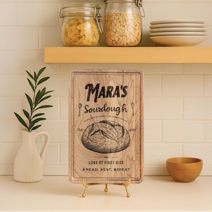 May include: A wooden cutting board with the text "Mara's Sourdough" and a bread illustration. The board is displayed on a gold stand. A white vase with greenery, a wooden bowl, and jars of food are in the background.