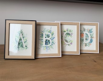 May include: Four framed art prints displaying the letters A, B, C, and D. Each letter is adorned with watercolour floral designs in green, blue, and pink hues. The frames are a mix of black and light wood.