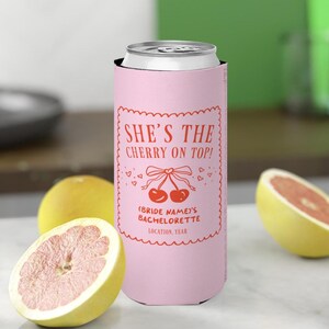 Könnte beinhalten: Ein rosa Getränkedosenkühler mit dem Text "SHE'S THE CHERRY ON TOP!" in Rot. Das Design umfasst eine Kirschgrafik und die Worte "(BRIDE NAME)'S BACHELORETTE LOCATION, YEAR". Der Kühler ist von halbierten Grapefruits und einer ganzen Grapefruit umgeben.
