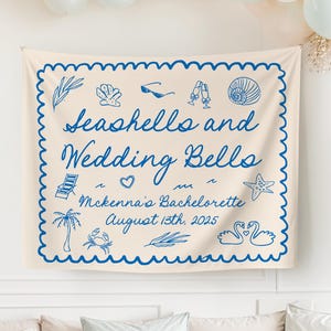 Pode incluir: Uma tapeçaria creme com bordas azuis onduladas e ilustrações de conchas, óculos de sol e taças de champanhe. O texto diz "Seashells and Wedding Bells" e "McKenna's Bachelorette August 13th, 2025". Balões e um sofá ao fundo.