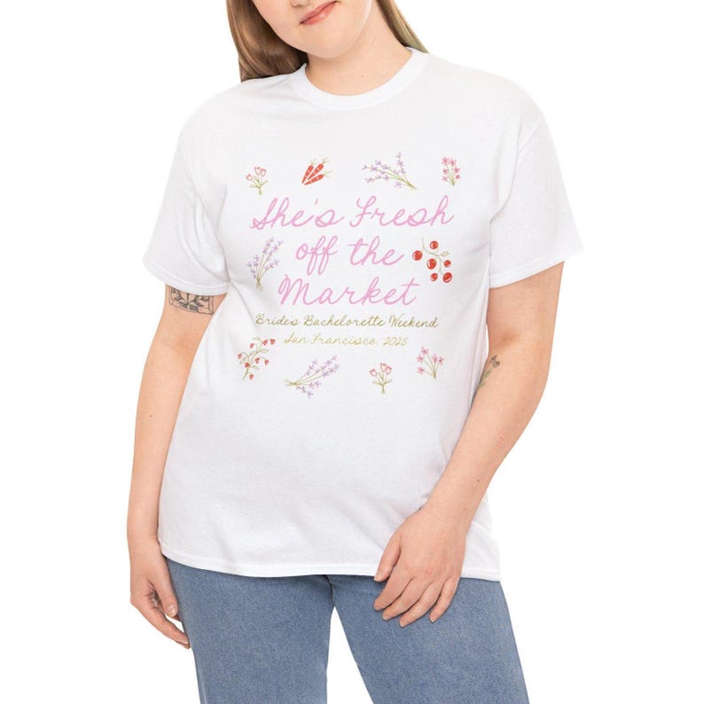 May include: White t-shirt with the text "She's Fresh off the Market" in pink script, decorated with floral illustrations. Below the text is "Bride's Bachelorette Weekend San Francisco 2025". The shirt is worn by a person.