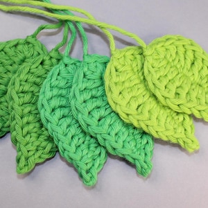 Puede incluir: Cinco hojas de ganchillo verdes, cada una con un tono de verde diferente, están conectadas entre sí por una cuerda blanca.