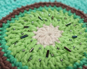May include: Close-up of a green, brown, and white crocheted circle with silver and blue beads. The center of the circle is white and the outer edge is green.