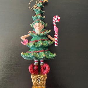 Puede incluir: Un adorno navideño caprichoso. El adorno tiene la forma de un elfo sonriente con un cuerpo de árbol verde, botas rojas y una base de maceta dorada. El elfo sostiene un bastón de caramelo y un guante rosa. Una estrella dorada corona el árbol.