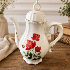 Peut inclure: Théière en céramique blanche avec un motif de roses. La théière présente un bec verseur incurvé, une anse ornée et un couvercle avec une boucle. Des roses rouges avec des feuilles vertes sont peintes sur le devant. Des bordures dorées ornent le couvercle et le bec. Sur une surface en bois.